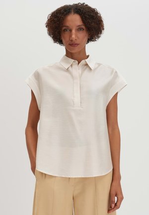 Femme aux cheveux bouclés portant un chemisier blanc à manches courtes avec col et un pantalon beige à jambes larges, debout devant un fond clair uni.