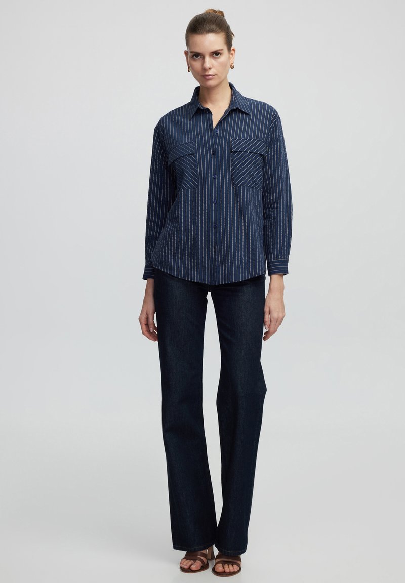 Marineblauwe langemouwshirt met dunne witte strepen, twee borstzakken, knopen aan de voorkant en puntkraag, gecombineerd met donkere jeans en bruine sandalen.