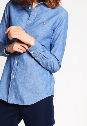 Persona con una camisa azul claro abotonada con un pequeño logo bordado en el pecho y pantalones oscuros, con las manos suavemente cruzadas a la altura de la cintura.