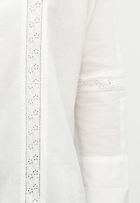 Λευκό ύφασμα με κατά μήκος και εγκάρσιες λεπτομέρειες δαντέλας με σχέδια λουλουδιών σε διαφανείς περιοχές υλικού.