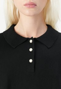 Juodas megztinis su apykakle ir keturiais perlų mygtukais. Audinys atrodo minkštas ir lygus. Detalus apykaklės vaizdas.