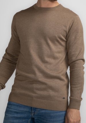 Mann trägt einen hellbraunen, gestrickten Pullover mit gerippten Bündchen und Saum, kombiniert mit blauen Jeans vor einem einfarbigen Hintergrund.