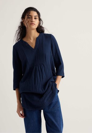 Hosszú hullámos hajú nő, aki texturált tengerészkék V-nyakú felsőt és kék farmert visel, egy egyszínű világos háttér előtt áll.