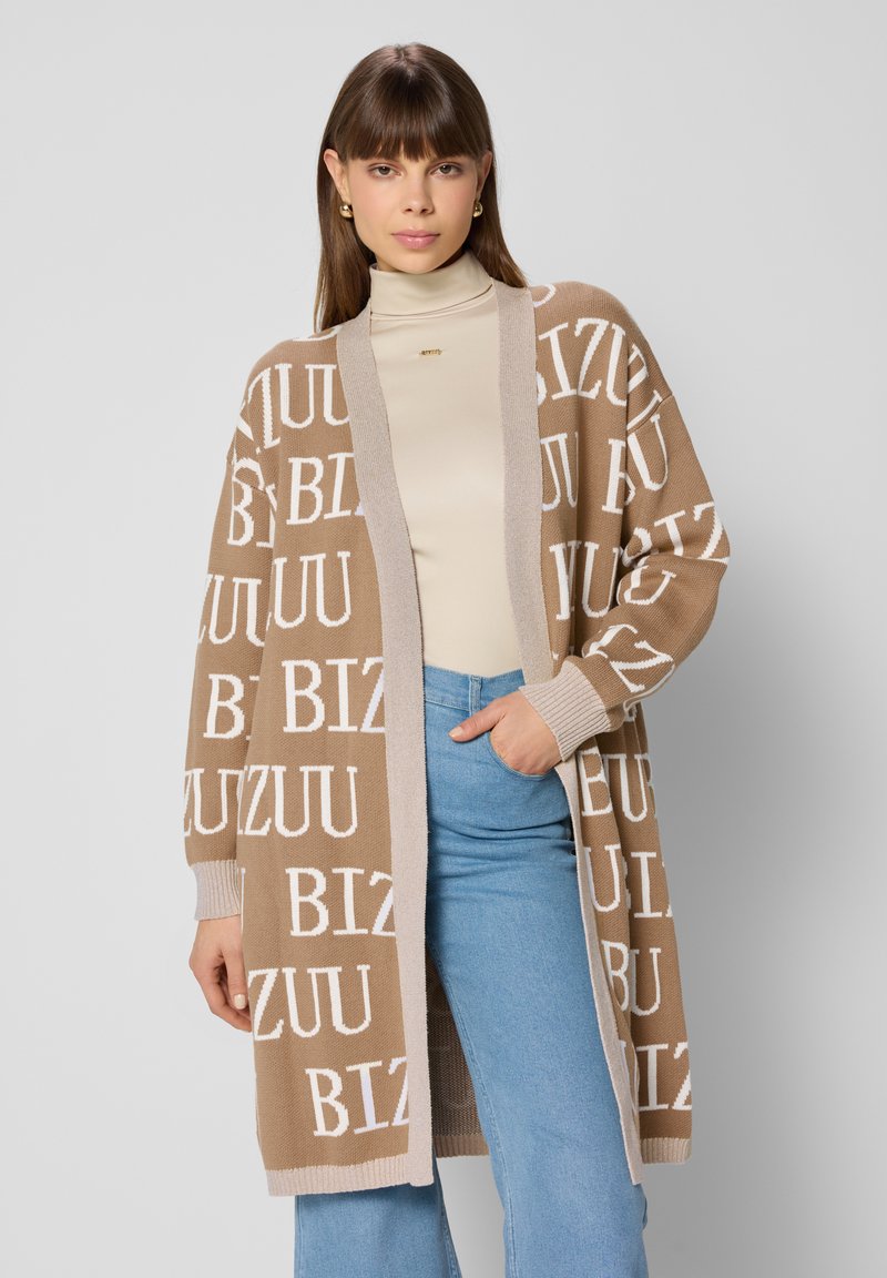 Losse bruine cardigan met grote witte tekst "BIZUU" verspreid over de stof, met een contrasterende beige rand. Gecombineerd met een beige coltrui.