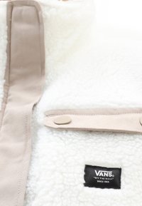 Hvid fleece jakke med beige detaljer; har et fremtrædende mærke, glat tekstur, trykknaplukning og et højt kravedesign.