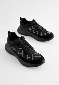 Black athletic shoes with a mesh and suede upper, featuring a crisscross pattern in white, and a thick, textured black sole.
