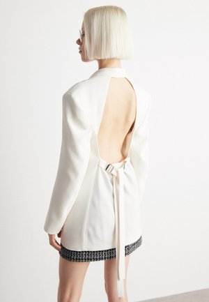 Blazer bianco con schiena aperta, dotato di chiusura a fiocco e maniche lunghe. L'orlo presenta un dettagliato accento nero texturizzato.