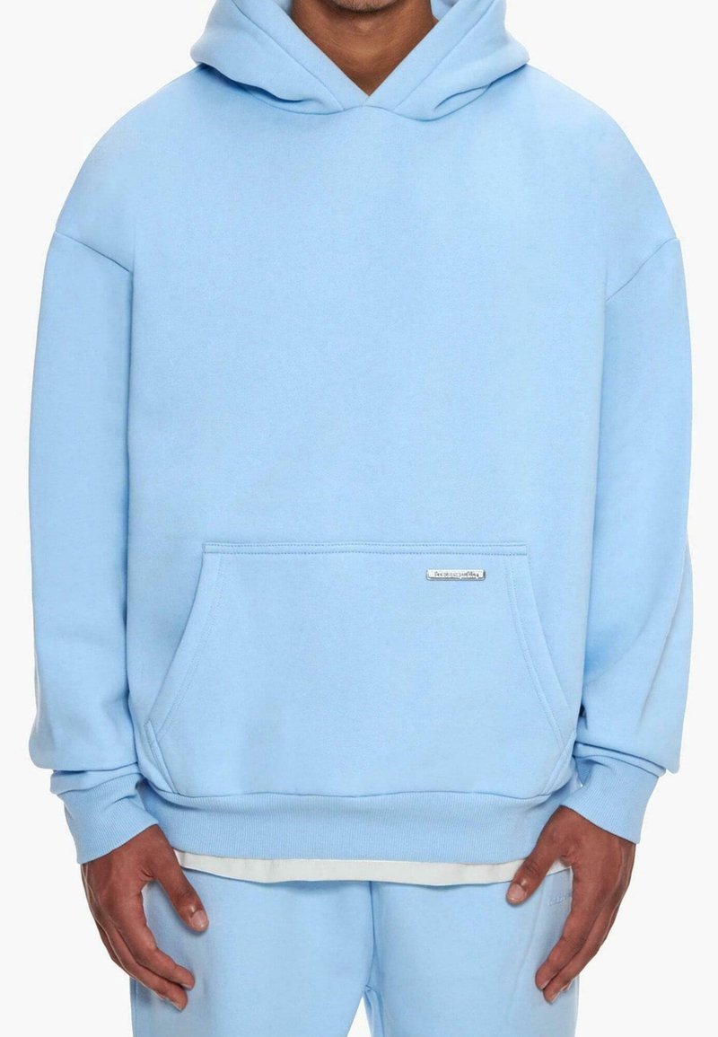 Homme portant un sweat à capuche oversize bleu clair avec une poche ventrale et un pantalon assorti bleu clair, les mains détendues le long du corps.