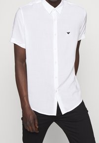 Man klädd i en kortärmad vit skjorta med knappar och svart broderad logotyp på bröstet, samt svarta byxor, står med handen i fickan.