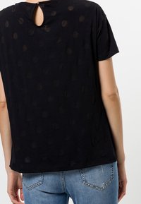 Top negro de manga corta con un sutil patrón de puntos, que presenta un escote en forma de lágrima en la parte posterior, hecho de una tela suave. Se lleva con jeans de mezclilla azules.