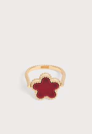 Bague en or avec des détails perlés présentant une pierre centrale rouge en forme de fleur sur un fond clair uni.