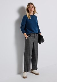 Pull bleu, pantalon taille haute à carreaux gris, baskets blanches avec des accents marron. Mannequin tenant une pochette noire, se tenant contre un fond blanc.