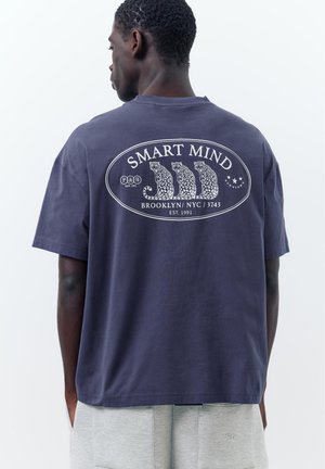 Muž, ktorý má na sebe tmavomodré tričko s tromi ilustráciami leopardov a textom „SMART MIND Brooklyn NYC 3743“ na chrbte.