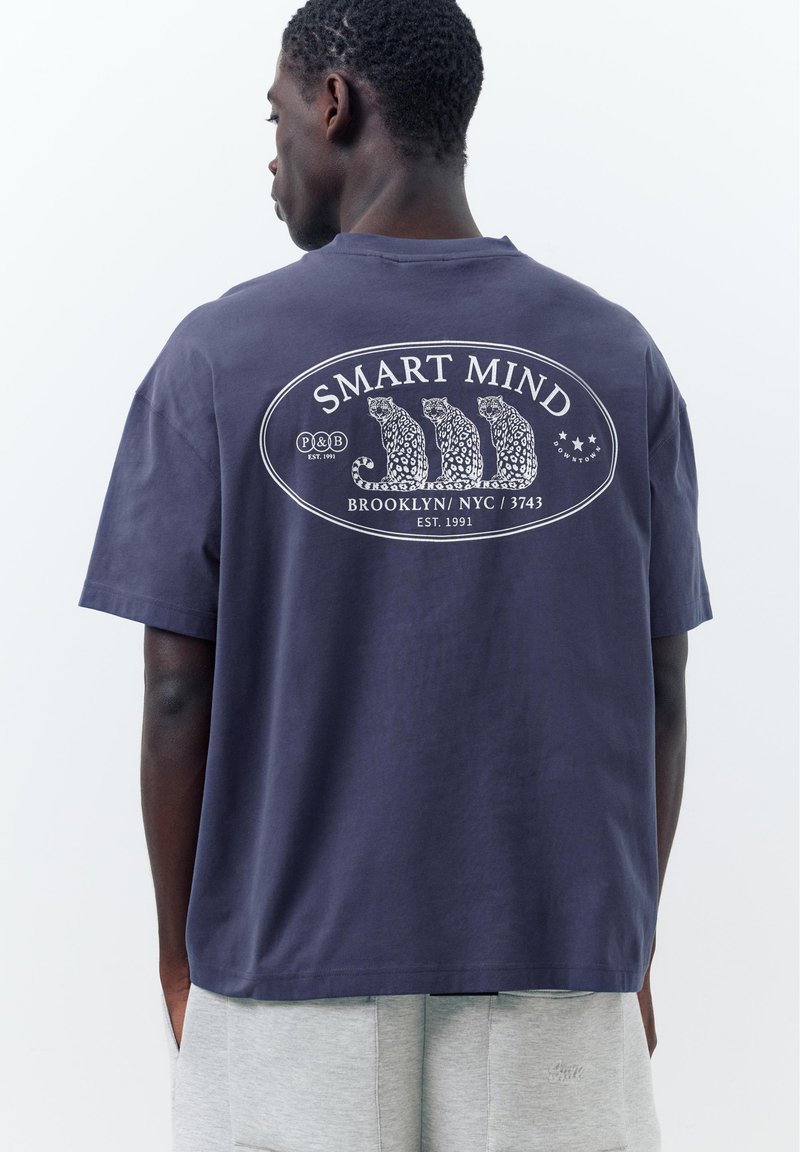 Homme portant un t-shirt bleu marine avec trois illustrations de léopard et le texte « SMART MIND Brooklyn NYC 3743 » dans le dos.