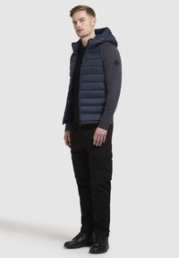 Gilet matelassé bleu marine avec capuche amovible, léger, texture matelassée, associé à une chemise à manches longues noire et un pantalon cargo, complété par des bottes noires.