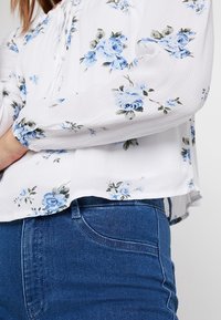 Floral blouse with white fabric featuring blue and green roses, long sleeves with gathered cuffs, paired with dark blue denim jeans.