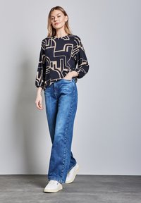 Haut à manches longues noir et beige avec un motif abstrait, associé à un jean large bleu et des baskets blanches sur un fond gris.