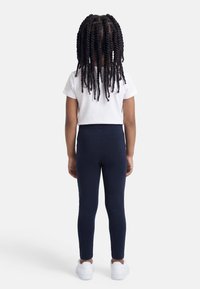 Παιδί με μακριά πλεγμένα κοτσίδια, φοράει λευκό μπλουζάκι, μπλε σκουρόχρωμο κολάν και λευκά αθλητικά παπούτσια, στέκεται με την πλάτη γυρισμένη σε λευκό φόντο.