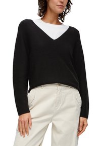 Schwarzer, gerippter Strickpullover mit V-Ausschnitt, langen Ärmeln und entspannter Passform, kombiniert mit hellbeigen Hosen mit seitlichen Taschen.