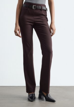 Femme portant un pantalon droit brun foncé avec une ceinture noire, des mocassins noirs à talons et un haut gris, debout sur un sol gris.