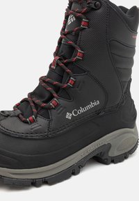 Botte d'hiver noire avec lacets rouges, tige en textile, bout renforcé et semelle en caoutchouc texturé. Présente le logo Columbia et un design étanche.