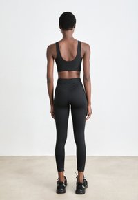 Femme de dos, portant un soutien-gorge de sport noir, des leggings taille haute et des baskets noires, devant un fond de mur blanc uni.