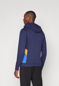 Sweatshirt azul-marinho com um tecido texturizado, possui capuz e detalhes laterais em azul e amarelo. Design casual e ajustado com punhos canelados.