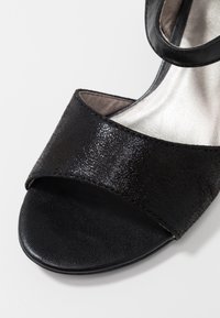 Svart sandal med ett slätt läderrem och texturerad ovandel med en glänsande finish; öppen tådesign och cirkulär sula.