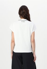 Balta īsām piedurknēm T-kreklu ar apaļu apkakli, kas izceļ melno akcentu. Uz aizmugures, tuvu kakla izgriezumam, redzams "KARL LAGERFELD" logotips.