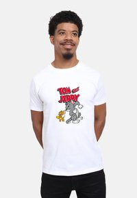 Bílý bavlněný tričko s potiskem kresleného seriálu Tom a Jerry. Design obsahuje výrazný červený text a barevné ilustrace postav.