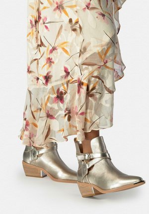 Botines metálicos brillantes con correas de hebilla combinados con un dobladillo de falda transparente con estampado floral en beige.