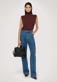 Burgunderfarbener ärmelloser Rollkragenpulli, hochgeschnittene blaue Jeans-Cargohose mit seitlichen Taschen, hält eine schwarze strukturierte Handtasche. Spitz zulaufende braune Schuhe.