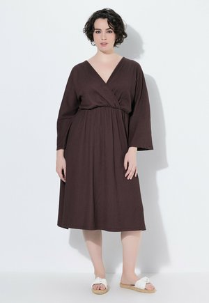 Kvinde med kort mørkt hår iført en brun midi-kjole med lange ærmer og hvide slip-on sandaler, stående mod en hvid baggrund.