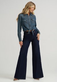 Denim shirt with a tie-front design, light blue color, featuring two chest pockets. Dark blue flared jeans with a high waist and stitched accents.