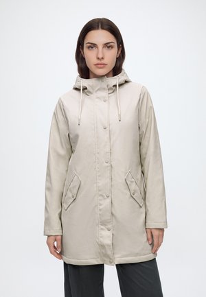 Mujer con chubasquero beige con capucha, botones a presión y bolsillos con tapeta, de pie frente a un fondo claro y liso.