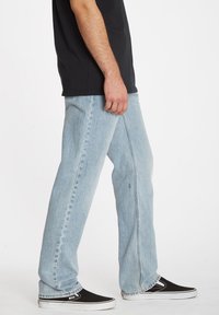 Vaqueros de pierna recta azul claro, hechos de denim, con costuras visibles y una cinturilla estándar, combinados con zapatillas de lona negras.