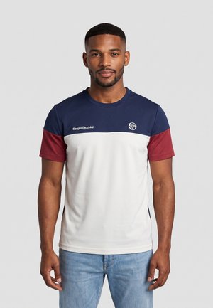 Uomo che indossa una camicia a maniche corte color-block in blu navy, bianco e bordeaux con il logo "Sergio Tacchini", abbinata a jeans azzurri.