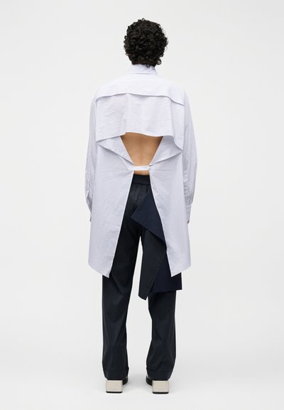 Person mit lockigem Haar trägt ein langärmeliges, hinten offenes gestreiftes Hemd, kombiniert mit Nadelstreifen-Hosen und Plateauschuhen, steht mit dem Rücken zur Kamera.