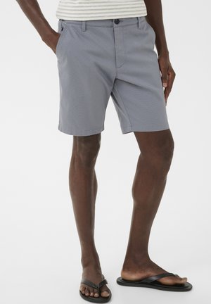 Mand iført lysegrå shorts, en stribet skjorte og sorte flip-flops, stående med hænderne i lommerne på en hvid baggrund.