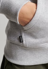 Sudadera gris con puños y dobladillo acanalados. Cuenta con un bolsillo frontal y una pequeña etiqueta negra "SUPERDRY" en el lateral. Textura de tela suave.
