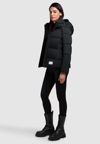 Sort pufferjakke med hætte, med lynlås, brystlomme og logo-detalje. Båret med sorte tights og ankelhælsko med snørebånd.