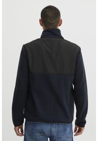 Giacca in pile blu navy con pannello superiore in nylon nero, colletto alto e finitura testurizzata. Presenta maniche lunghe e una vestibilità rilassata.