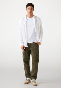 Witte linnen button-up shirt over een plain wit t-shirt, gecombineerd met olijfgroene cargobroek met zijzakken en beige sneakers.