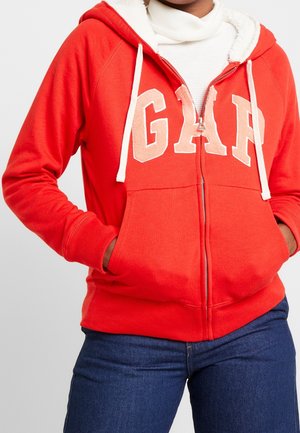 Person iført en rød hættetrøje med hvid fleeceforing og "GAP" bogstaver på brystet, kombineret med blå jeans og en hvid top.