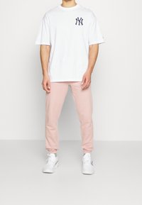 Biała bawełniana koszulka z granatowym logo na piersi, połączona z różowymi dresami z polaru i białymi sneakersami. Prosty design, luźny krój, bez wzorów.