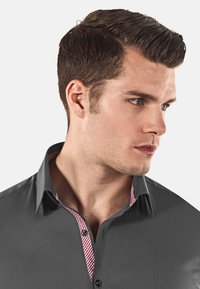 Camicia grigia con colletto button-down, caratterizzata da un interno a scacchi rosso e bianco. Tessuto liscio con leggero lucido e bottoni a contrasto.