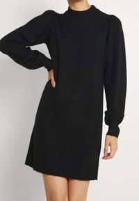 Robe en tricot noire à manches longues avec des épaules bouffantes et des poignets côtelés, portée par une personne avec une main sur la hanche et l'autre pendant.