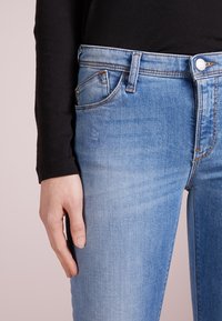 Bleka blå denimjeans med femficksdesign, ljusa slitningar och en klassisk knäppning i midjan. Svart långärmad topp delvis synlig.