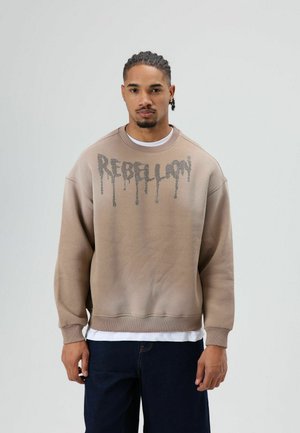 Bruine sweatshirt met een verloopontwerp, met daarop "REBELION" in druppelende letters. Geribbelde manchetten en zoom, gedragen over een wit shirt.
