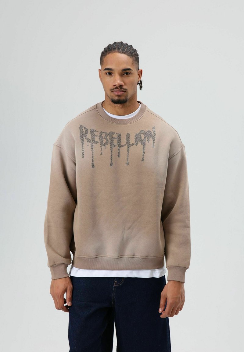 Bruine sweatshirt met een verloopontwerp, met daarop "REBELION" in druppelende letters. Geribbelde manchetten en zoom, gedragen over een wit shirt.